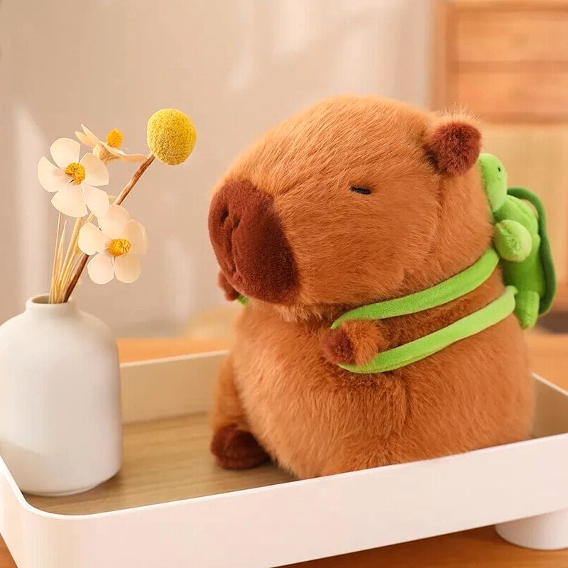 Capybara Dreams plush backpack toy in soft brown fabric with a cute green turtle design, sitting in a white decorative tray next to a small white vase with yellow and white flowers. The adorable capybara plush has a sleepy embroidered face and a functional mini backpack.