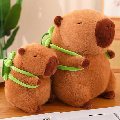 Two Capybara Dreams plush backpack toys in soft brown fabric with cute green turtle designs, showcasing different sizes. The larger and smaller capybara plushies sit on a wooden surface next to an open book, each with a sleepy embroidered face and a mini backpack.