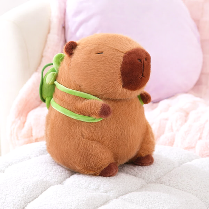 Capybara Dreams plush backpack toy, soft brown fabric, featuring a cute green turtle design on its back, sitting on a white textured surface.