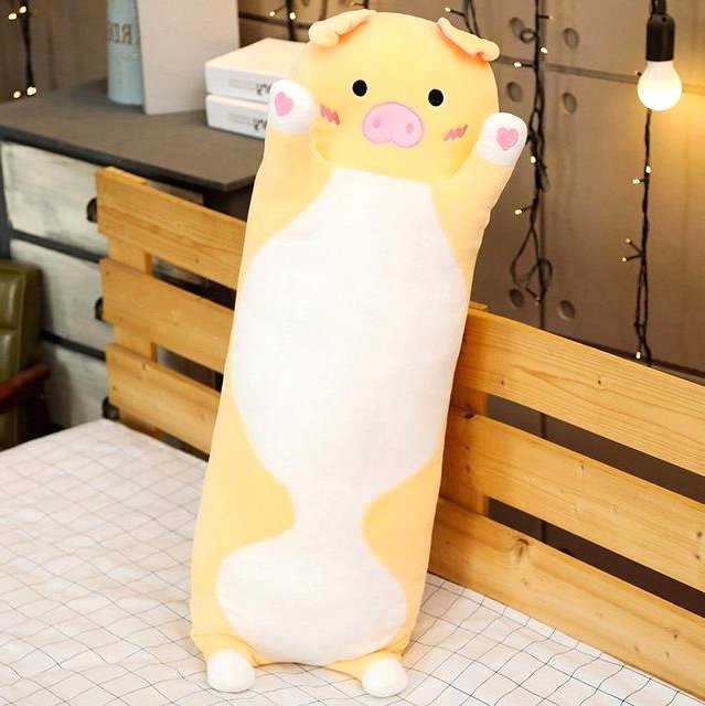 A long yellow pig plush toy with pink cheeks and paw pads standing upright on a bed. In the background, there is a wooden headboard, string lights, and shelves. The plush toy has a soft fabric and a huggable design.