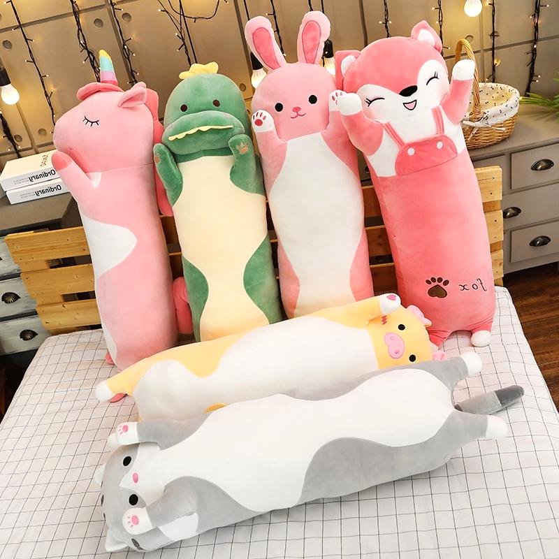 Cuddle Haven collection of long plush toys, including a pink fox, pink bunny, green dinosaur, pink unicorn, gray cat, yellow cat, displayed together on a bed. Showcases various soft fabric animal plushies.