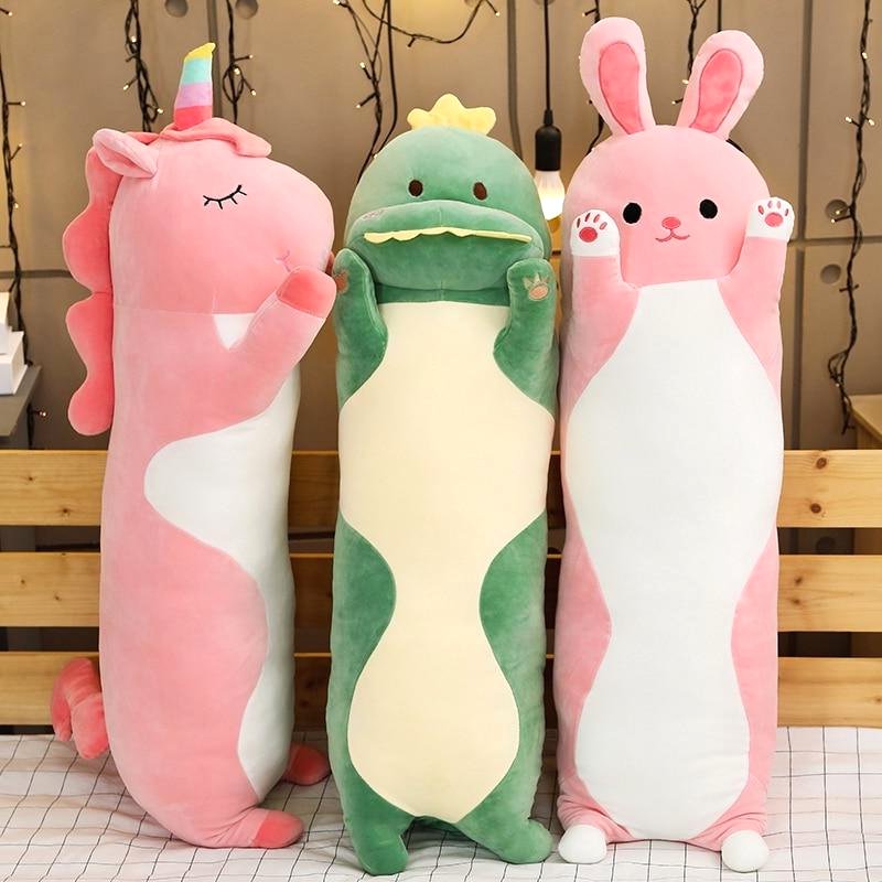Three Cuddle Haven long plush toys standing on a bed. From left to right: a pink bunny plush, a green dinosaur plush, and a pink unicorn plush. All feature soft fabric a huggable design, ideal as cuddle buddies or decorative plush.