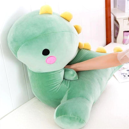 A hand gently pressing into the soft, green dinosaur plush toy, demonstrating its squishy texture. The plush toy has a round head, small yellow spikes, and a cute embroidered face.