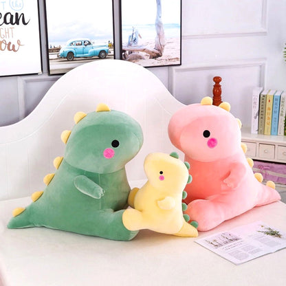 A trio of soft fabric dinosaur plush toys, including a large pink, a small yellow, and a large green dino, arranged on a white bedspread with books and framed art in the background.